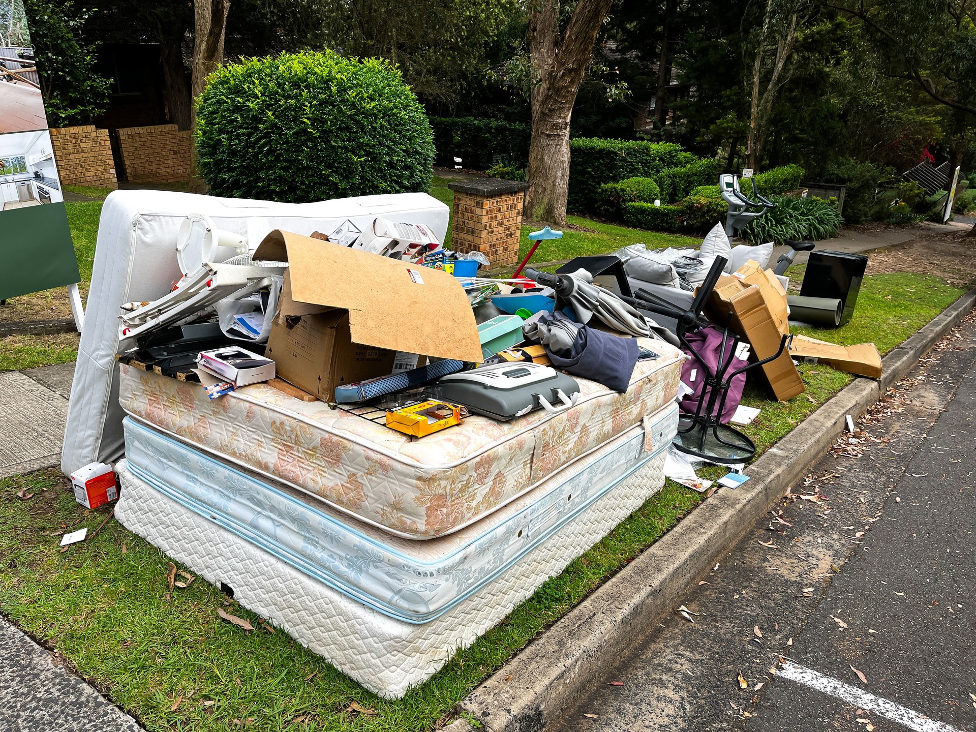 A pile of rubbish on the curb for council collection, including cardboard boxes, a couch with pillows, stacked mattresses, and coat hangers