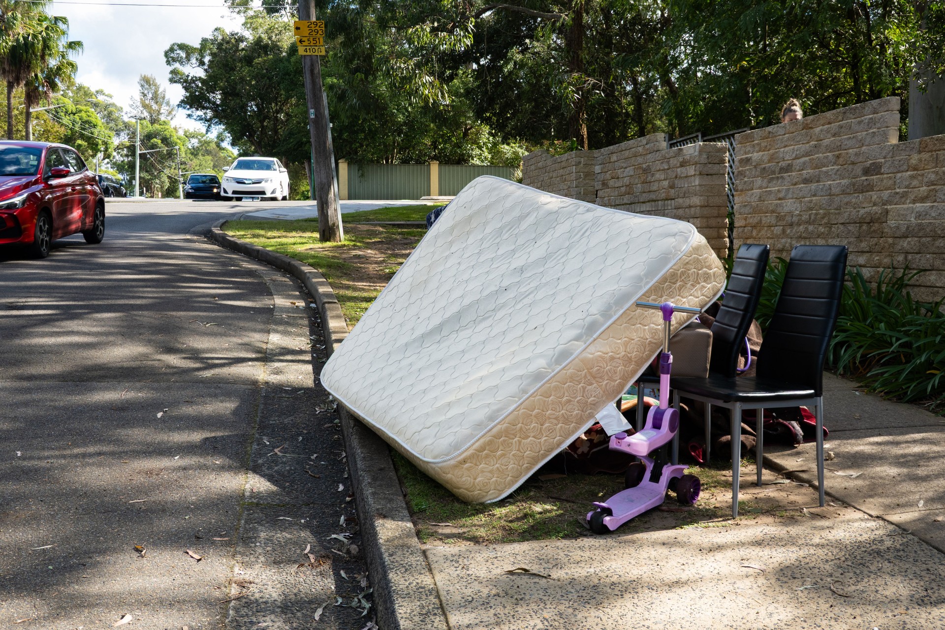A pile of rubbish on the curb for council collection, including cardboard boxes, a chair, stacked mattresses, and other dumped material